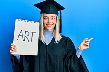 Beautiful blonde woman wearing graduation cap and ceremony robe holding art notebook smiling happy...