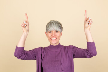 Stylish woman in purple casual on beige background happy excited cheerful point up with index finger