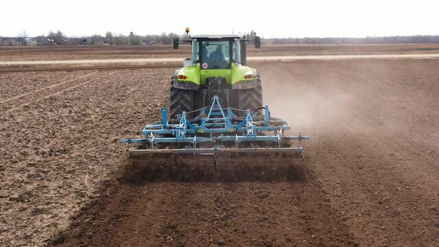 Tractor with harrow system plowing ground on cultivated farm field, pillar of dust trails behind, preparing soil for planting new crop, agriculture concept, top view.