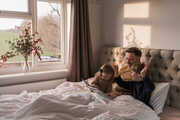 Obraz na płótnie Canvas Happy dad reads a book to children lying on the bed. Dad, son and daughter. Joy. Day.