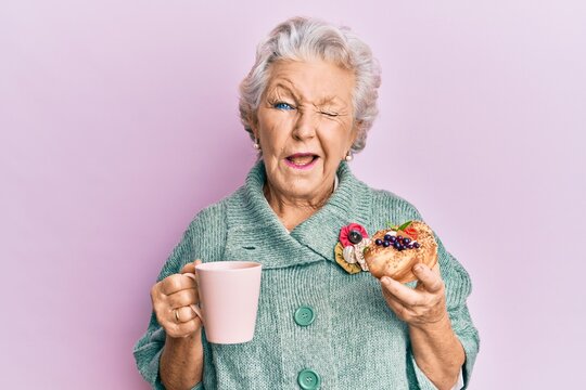 Senior grey-haired woman drinking a cup of coffee and eating bun winking looking at the camera with sexy expression, cheerful and happy face.