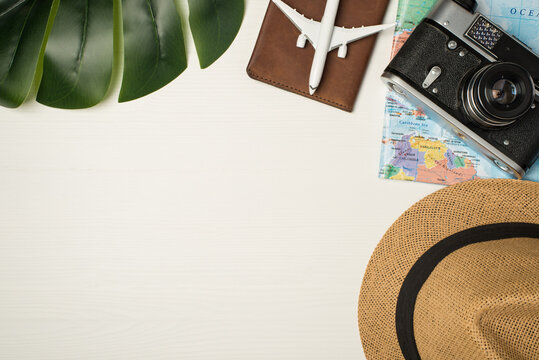 Top view photo of green palm leaf plane model on passport cover sunhat camera and map on wooden white background with copyspace