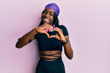 Young african american woman wearing gym clothes and using headphones smiling in love doing heart symbol shape with hands. romantic concept.