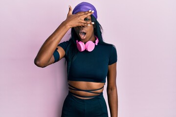Young african american woman wearing gym clothes and using headphones peeking in shock covering face and eyes with hand, looking through fingers with embarrassed expression.