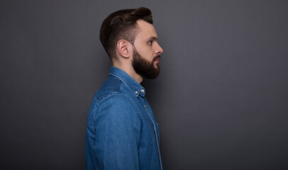Obraz premium Close up side view photo of handsome bearded modern confident man in jeans shirt on the gray background