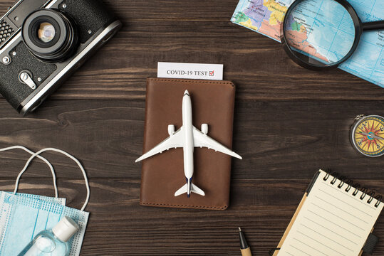 Top view photo of plane model on leather passport cover with covid test camera map magnifier compass notebook pen medical masks and sanitizer on isolated wooden table background