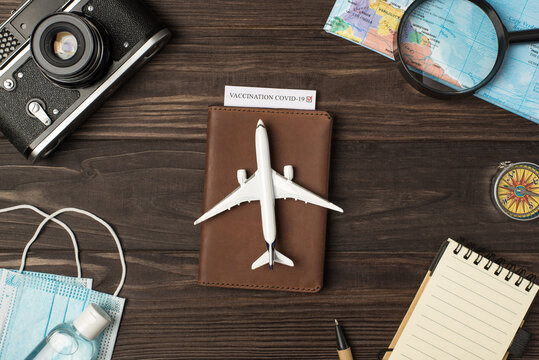 Top view photo of plane model on leather passport cover with vaccination certificate camera map magnifier compass notebook pen medical masks and sanitizer on isolated wooden table background