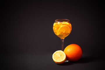 Glass of white wine sangria with orange and lemon, orange and half of lemon near. Dark background.
