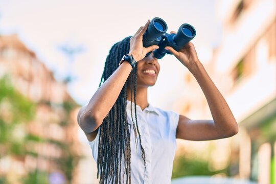 Young african american woman smiling happy looking for new opportunity using binoculars at the city.