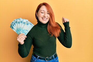 Beautiful redhead woman holding 100 brazilian real banknotes screaming proud, celebrating victory...