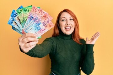 Beautiful redhead woman holding swiss franc banknotes celebrating achievement with happy smile and winner expression with raised hand