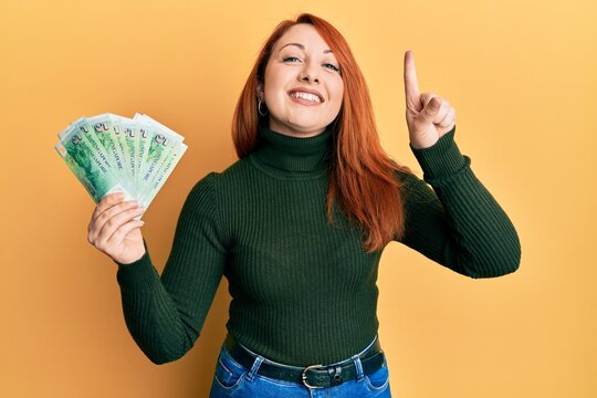 Beautiful redhead woman holding 5 singapore dollars banknotes smiling with an idea or question pointing finger with happy face, number one