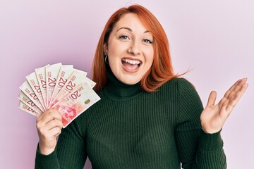 Beautiful redhead woman holding 20 israel shekels banknotes celebrating achievement with happy smile and winner expression with raised hand