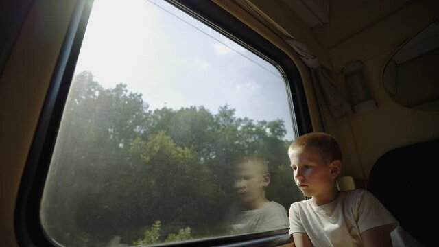 Boy is traveling by train. Kid looks out the train window. Thoughtful boy in a train compartment. The child dreams of a fun summer vacation. Long-distance family trip. End of lockdown. Tourist trip.
