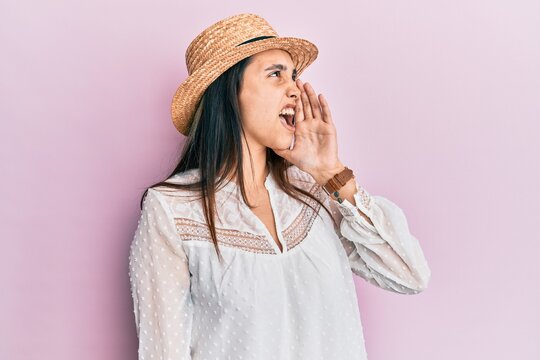 Young hispanic woman wearing summer hat shouting and screaming loud to side with hand on mouth. communication concept.