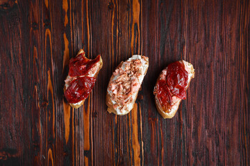 three bruschetta sandwiches on baguette bread with cottage cheese, sun-dried tomatoes and red fish on a dark wooden background, top view, copy space. Traditional italian snack for wine - grilled tapas