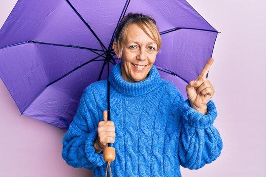Middle age blonde woman holding purple umbrella smiling happy pointing with hand and finger to the side