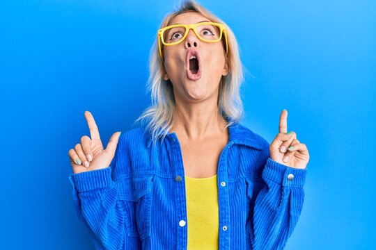 Young blonde girl wearing casual clothes and glasses amazed and surprised looking up and pointing with fingers and raised arms.