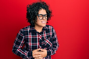Young hispanic woman with curly hair wearing casual clothes and glasses with hand on stomach because nausea, painful disease feeling unwell. ache concept.