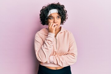 Young hispanic woman with curly hair wearing sportswear looking stressed and nervous with hands on mouth biting nails. anxiety problem.
