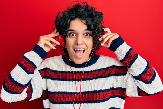 Young hispanic woman with curly hair listening to music using headphones celebrating crazy and amazed for success with open eyes screaming excited.