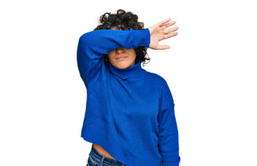 Young hispanic woman with curly hair wearing turtleneck sweater covering eyes with arm, looking serious and sad. sightless, hiding and rejection concept