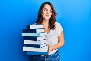 Young caucasian woman holding a pile of books smiling looking to the side and staring away thinking.