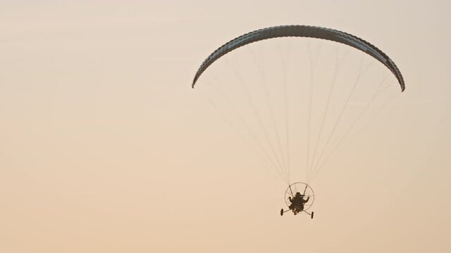 Paramotor flying away from camera. Parachute against sunset sky. Tandem motor powered paragliding at twilight. 