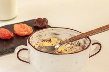 Muesli in white bowl, dried fruit on black cutting board and a bottle of milk on white background. Breakfast concept. Stock photo.