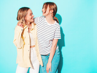 Two young beautiful blond smiling hipster female in trendy summer clothes. Sexy carefree women posing near blue wall in studio. Trendy and positive models having fun