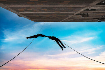 Two person silhouettes, dancing in the air, tied by the ropes on the roof. Extreme rope dance on the wall of a tall building on the sunset. 
