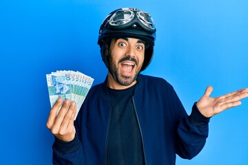 Young hispanic man wearing motorcycle helmet holding hong kong dollars celebrating achievement with happy smile and winner expression with raised hand