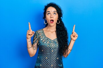 Young woman wearing bindi and traditional kurta dress amazed and surprised looking up and pointing with fingers and raised arms.