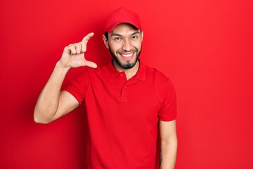 Hispanic man with beard wearing delivery uniform and cap smiling and confident gesturing with hand doing small size sign with fingers looking and the camera. measure concept. © Krakenimages.com