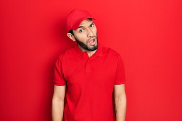 Hispanic man with beard wearing delivery uniform and cap in shock face, looking skeptical and sarcastic, surprised with open mouth