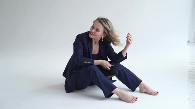 woman in blue suit sits barefoot on in white photo studio and holds heeled shoes