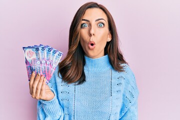 Young brunette woman holding hong kong 10 dollars banknotes scared and amazed with open mouth for...