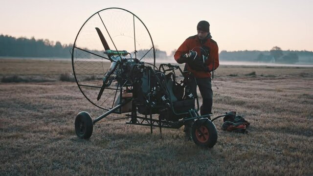 Man standing at paramotor, checking and preparing safety gear for customer. Tandem motor powered paragliding at twilight. 