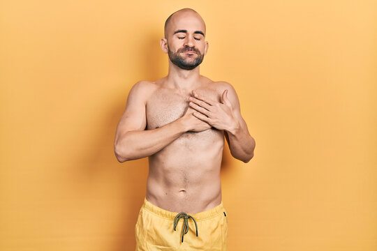 Young bald man wearing swimwear smiling with hands on chest with closed eyes and grateful gesture on face. health concept.