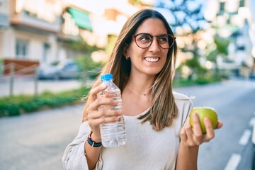 Young caucasian woman smiling happy holding green apple and bottle of water at the city.