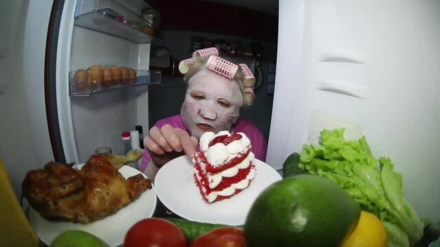 A woman wearing a cosmetic mask looks in the refrigerator for food. Makes a choice: smoked chicken or fruit with vegetables, or cake on a plate.