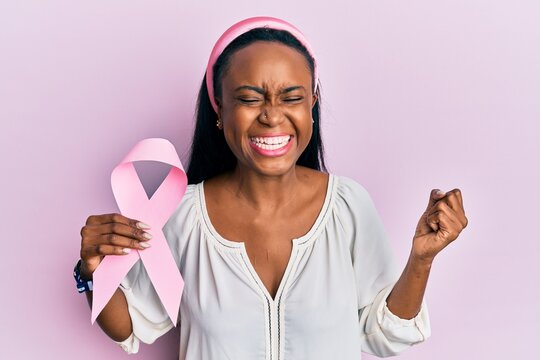 Young african woman holding pink cancer ribbon screaming proud, celebrating victory and success very excited with raised arm