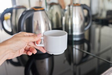 White coffee cup in hand close-up, stainless steel electric kettle in mirror background