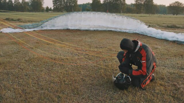 Pilot kneeling on grass and checking safety helmet. Unpacked parachute in background. Tandem motor powered paragliding at twilight. 