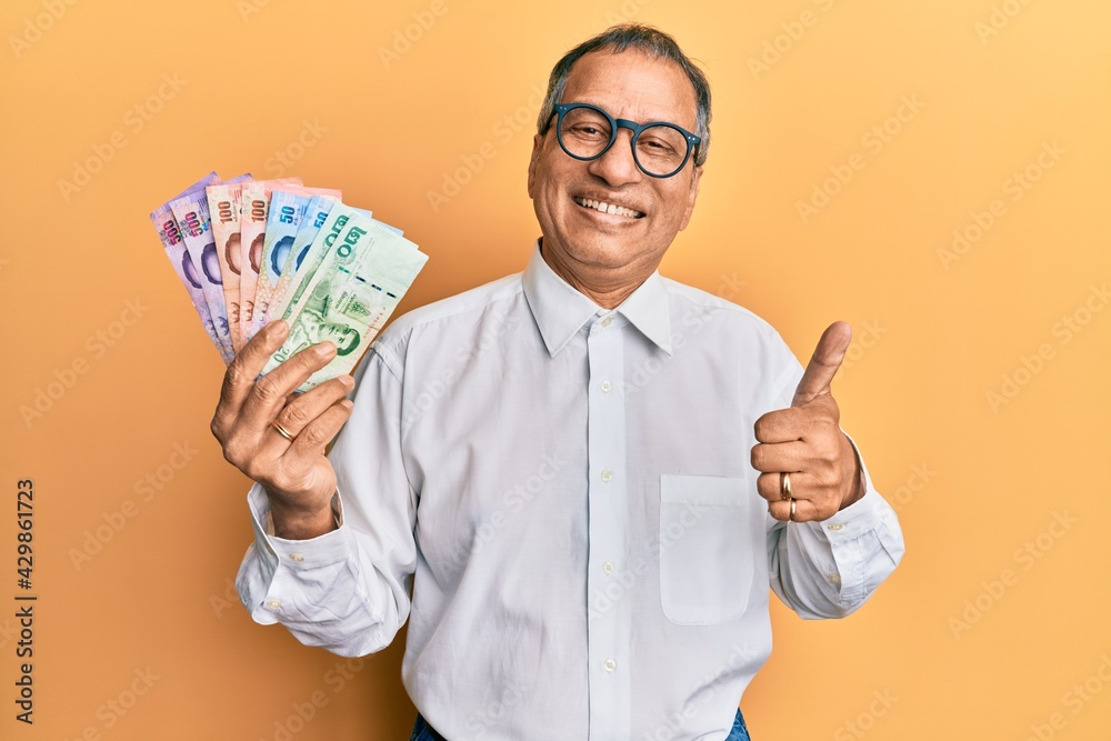 Sticker middle age indian man holding thai baht banknotes smiling happy and positive, thumb up doing excelle