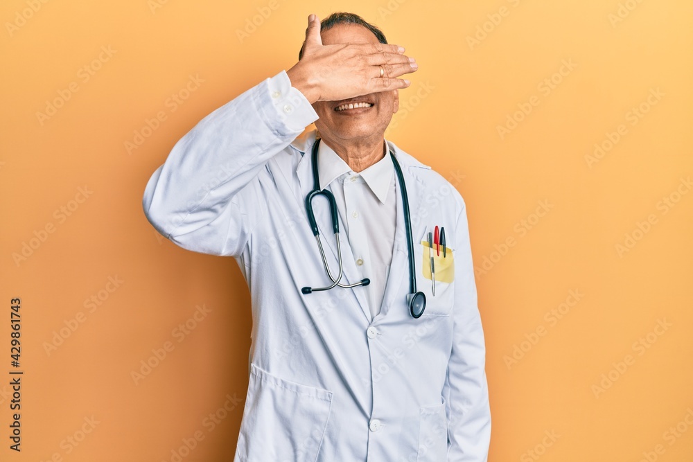 Sticker Middle age indian man wearing doctor coat and stethoscope smiling and laughing with hand on face covering eyes for surprise. blind concept.