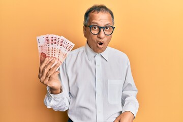 Middle age indian man holding 100 new taiwan dollars banknotes scared and amazed with open mouth for surprise, disbelief face