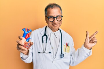 Middle age indian man wearing doctor uniform holding heart smiling happy pointing with hand and finger to the side