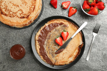 Delicious thin pancakes served with chocolate paste and strawberries on grey table, flat lay