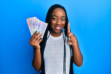 Young african american woman holding swedish krona banknotes smiling with an idea or question pointing finger with happy face, number one